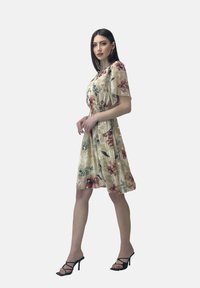 Vestido de estampado floral en crema, con mangas cortas y cintura ajustada. Combinado con tacones negros con tiras, mostrando un ajuste relajado.