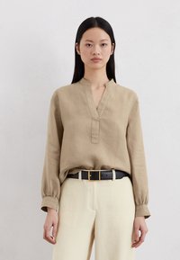 Beige Leinenhemd mit Mandarin-Kragen, langen Ärmeln und knöpfbarer Vorderseite. Tragen zu hoch geschnittenen cremefarbenen Hosen und einem schwarzen Gürtel.