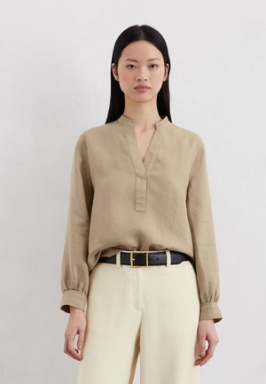 Chemise en lin beige avec col mandarin, manches longues et patte de boutonnage. Portée avec un pantalon crème taille haute et une ceinture noire.