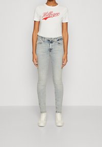 Camiseta blanca con el logo rojo de "Hilfiger", combinada con jeans ajustados de color azul claro y zapatillas blancas. Diseño simple y entallado; apariencia casual.