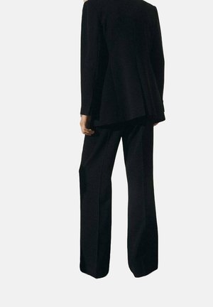 Personne portant un costume noir ample avec un long blazer et un pantalon à jambes larges, debout dos à la caméra.