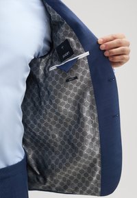Niebieska marynarka z wzorzystą srebrną podszewką. Wewnętrzna metka zawiera napis "JOOP!" oraz "SLIM FIT." Posiada jedną wewnętrzną kieszeń i kontrastujące faktury.