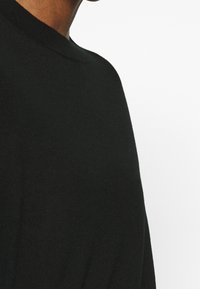 Närbild av en person som bär en svart rundhalsad tröja mot en vit bakgrund, som visar nacken och övre delen av torso.