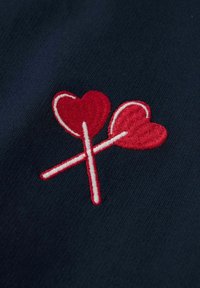 Corazones rojos bordados sobre palos cruzados contra un fondo azul marino oscuro. Presenta formas curvas y un acabado suave y texturizado.