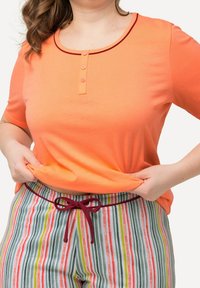 Top arancione a maniche corte con scollatura rotonda e pattina con tre bottoni, abbinato a pantaloni a righe multicolori con vita legata in bordeaux.