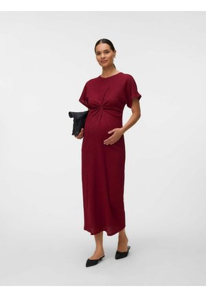 Gravid kvinde iført en rød midi kjole og sorte flade sko, der holder en sort clutch, stående mod en ensartet lys baggrund.