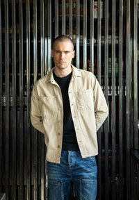 Beige knoopshirt over een zwart T-shirt, gecombineerd met blauwe spijkerbroek. Staand tegen een achtergrond van verticale metalen latten.