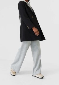 Zwarte double-breasted jas met metalen knopen, gecombineerd met lichtgrijze wijde sweatpants en beige sportschoenen.