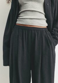 Person wearing loose black pants with elastic waist, gray ribbed cropped top, and black open-button shirt with one hand in pocket.