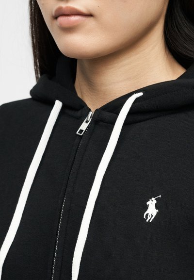 Κοντινό πλάνο ατόμου που φοράει μαύρο φούτερ με φερμουάρ, λευκά κορδόνια και μικρό λευκό κεντητό έμβλημα παίκτη πόλο στο στήθος.