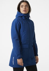 Blue waterproof parka with a high collar, hood, two front pockets, and logo detailing. Features a smooth texture and adjustable elements.