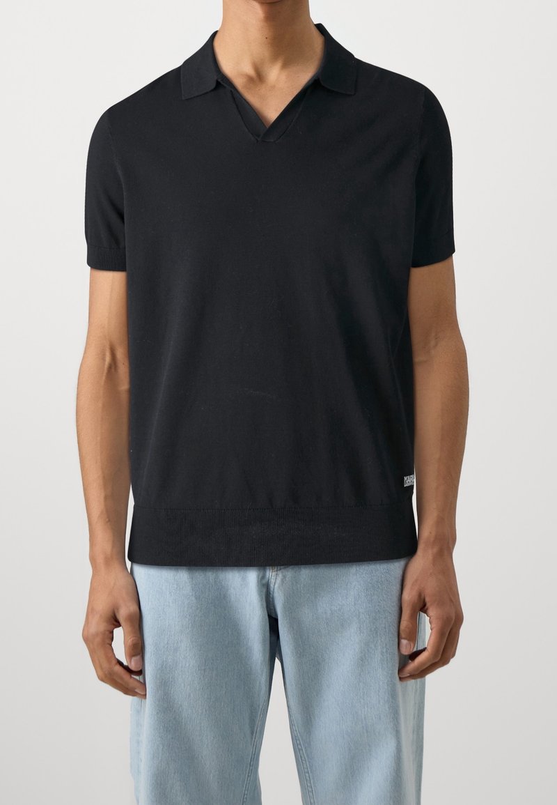 Homme portant un polo noir à manches courtes avec un col en V et un jean bleu clair, debout devant un fond uni.