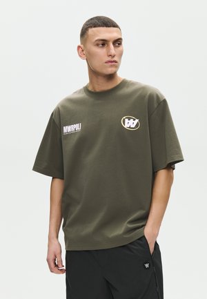 Ung mand med kort hår iført en olivengrøn oversize T-shirt og sorte bukser, stående med den ene hånd i lommen, kigger mod højre.