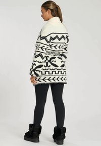 Stickad kofta i krämfärg med svarta geometriska mönster, sjalkrage, långa ärmar och fransad nederkant. Kombineras med svarta leggings och stövlar med päls.