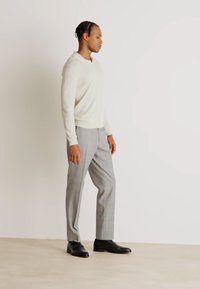 Lichtbeige gebreide trui met kraag, gecombineerd met grijs geruite broek. Zwarte leren schoenen maken de outfit compleet. Zijaanzicht.
