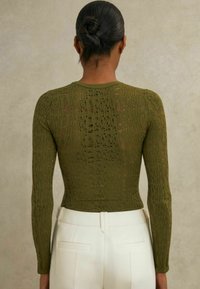 Olíva zöld, hosszú ujjú felső, bonyolult nyitott kötésű dizájnnal, kerek nyakkivágással és testhezálló szabással, fehér nadrággal párosítva.
