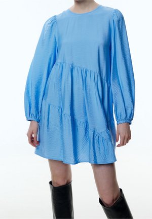 Kvinde iført en lyseblå, langærmet, lagdelt minikjole med tekstureret stof og sorte knæhøje støvler, stående mod en hvid baggrund.