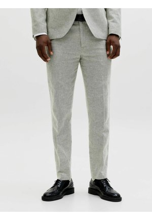 Mężczyzna w jasnoszarych spodniach od garnituru, dopasowanej marynarce, białej koszuli, czarnym pasku i czarnych skórzanych eleganckich butach, stojący na białym tle.