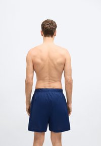 Άνδρας που στέκεται με την πλάτη, χωρίς πουκάμισο, φορώντας σκούρα μπλε μαγιό, σε απλό φωτεινό φόντο.