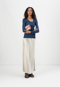 Top de manga longa canelado em azul-marinho, saia maxi em cetim bege e sandálias pretas. Modelo segura uma clutch rosa. Design simples e ajustado.