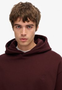 Sudadera con capucha color burdeos hecha de tela suave, con un bolsillo frontal tipo canguro y un sutil logo en el pecho en un tono a juego.