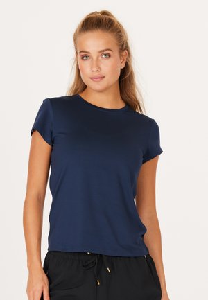 Lyshåret kvinde med hestehale iført en mørkeblå kortærmet T-shirt og sorte bukser med snøre, stående mod en ensfarvet lys baggrund.
