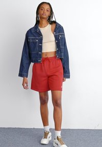 Veste en jean au design court, de couleur bleu clair ; haut moulant beige ; short de sport rouge avec une taille élastique ; baskets blanches avec des accents.