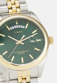Montre Timex avec un cadran vert, des accents dorés, fonction jour-date, boîtier en acier inoxydable et bracelet en métal argenté et doré.