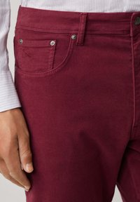 Bordeauxkleurige jeans met een gladde textuur, voorzien van een voorzak met metalen hardware en gestikte details op de zakken en de tailleband.