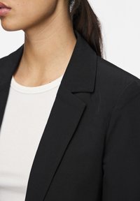 Černý blazer se strukturovaným klopem, nošený přes bílé tričko s žebrováním. Tkanina vypadá hladce, vykazuje minimální texturu a žádné viditelné vzory.