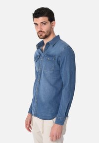 Camicia di denim blu medio con colletto button-down, due tasche sul petto e una vestibilità affusolata. Maniche lunghe con polsini a bottoni.