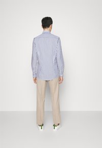 Chemise rayée à manches longues en blanc et bleu marine, avec des poignets boutonnés, associée à un pantalon beige clair et des chaussures blanches avec des accents verts.