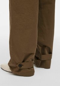 Bruine wijde broek gemaakt van gestructureerde stof, met knoopboorden en een ontspannen pasvorm. Eindigt net boven een witte schoen.