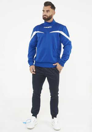 Uomo con barba che indossa una giacca sportiva blu e bianca, pantaloni sportivi neri e sneakers bianchi, in piedi con una mano in tasca.