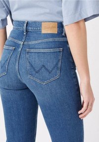 Jeans de mezclilla azul con cintura alta, que presentan una etiqueta de cuero marcada con "Wrangler" y dos bolsillos traseros con acentos de costura en zigzag.