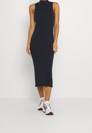 Femme portant une robe midi noire côtelée sans manches, associée à des baskets blanches de sport, debout devant un fond blanc uni.