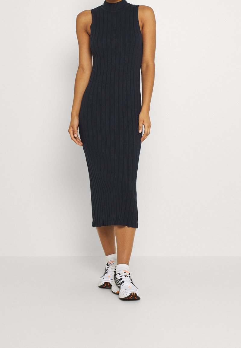 Femme portant une robe midi noire côtelée sans manches, associée à des baskets blanches de sport, debout devant un fond blanc uni.