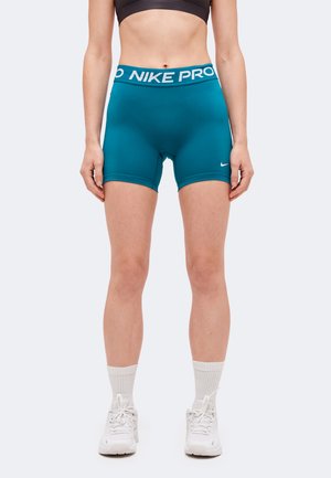 Modèle portant un short ajusté Nike Pro bleu canard, une brassière de sport noire, des chaussettes blanches mi-mollet et des baskets blanches sur un fond uni.