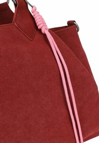 Suède handtas in dieprood met een gestructureerde afwerking. Voorzien van roze gevlochten koorden en metalen hardware. Gelaagd ontwerp met een open bovenkant.