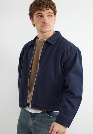 Młody mężczyzna z kręconymi włosami, ubrany w granatową kurtkę na suwak, na brązowym swetrze i niebieskich dżinsach, patrzy lekko w bok.