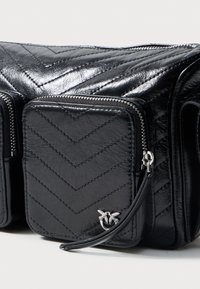 Bolsa de couro preto acolchoada com três compartimentos com fecho, costura em ziguezague e um pequeno emblema de pássaro prateado no bolso da frente.