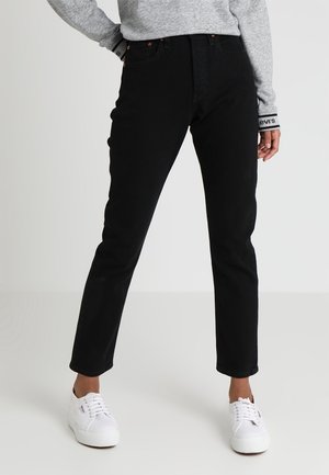 Person trägt schwarze Jeans mit hoher Taille und geradem Bein, grauen Pullover mit Levi’s-Logo am Ärmel und weiße Schnürsneaker und steht vor einem einfarbigen Hintergrund.