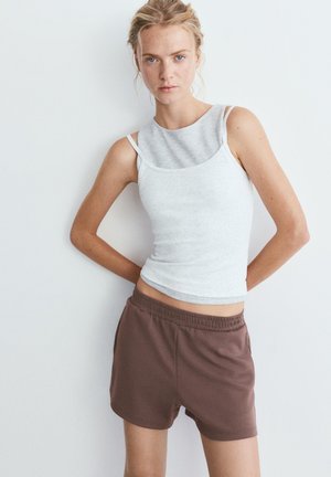 Jeune femme aux cheveux blonds portant un débardeur gris clair à plusieurs couches et un short marron à taille élastique, debout contre un mur blanc.