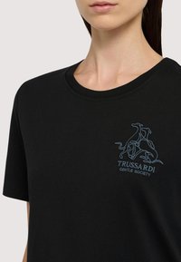 Czarny bawełniany T-shirt z okrągłym dekoltem. Posiada haftowane jasnoniebieskie logo przedstawiające stylizowane chartki i napis „TRUSSARDI GENTLE SOCIETY.”