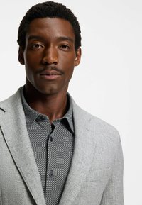 Grijze wollen blazer over een zwarte geblokte shirt met knopen. De stof lijkt textuur te hebben met een klassieke kraag en een getailleerde pasvorm.