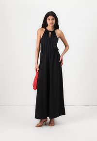 Vestido maxi preto com um decote halter e detalhe em buraco, cintura ajustada, tecido fluido, e combinado com uma bolsa vermelha e saltos com tiras.