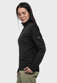 Schwarzer Quarter-Zip-Pullover mit gerippter Textur, langen Ärmeln und engem Kragen. Mit Logo auf der Brust und dem Ärmel. Dazu eine hellgrüne Hose.