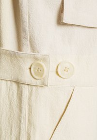 Beige látka s texturovaným povrchem má dva velké, kulaté, krémové knoflíky a detail klopového límce poblíž výstřihu.