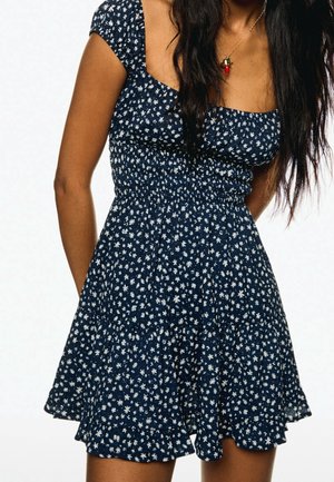 Mujer con un vestido azul marino con estampados florales blancos y un escote cuadrado, combinado con un collar colgante en forma de chile.