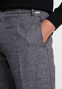 Grå och svart rutiga byxor med en sidficka och en etikett på midjan. Tyget har en texturerad, vävd yta.